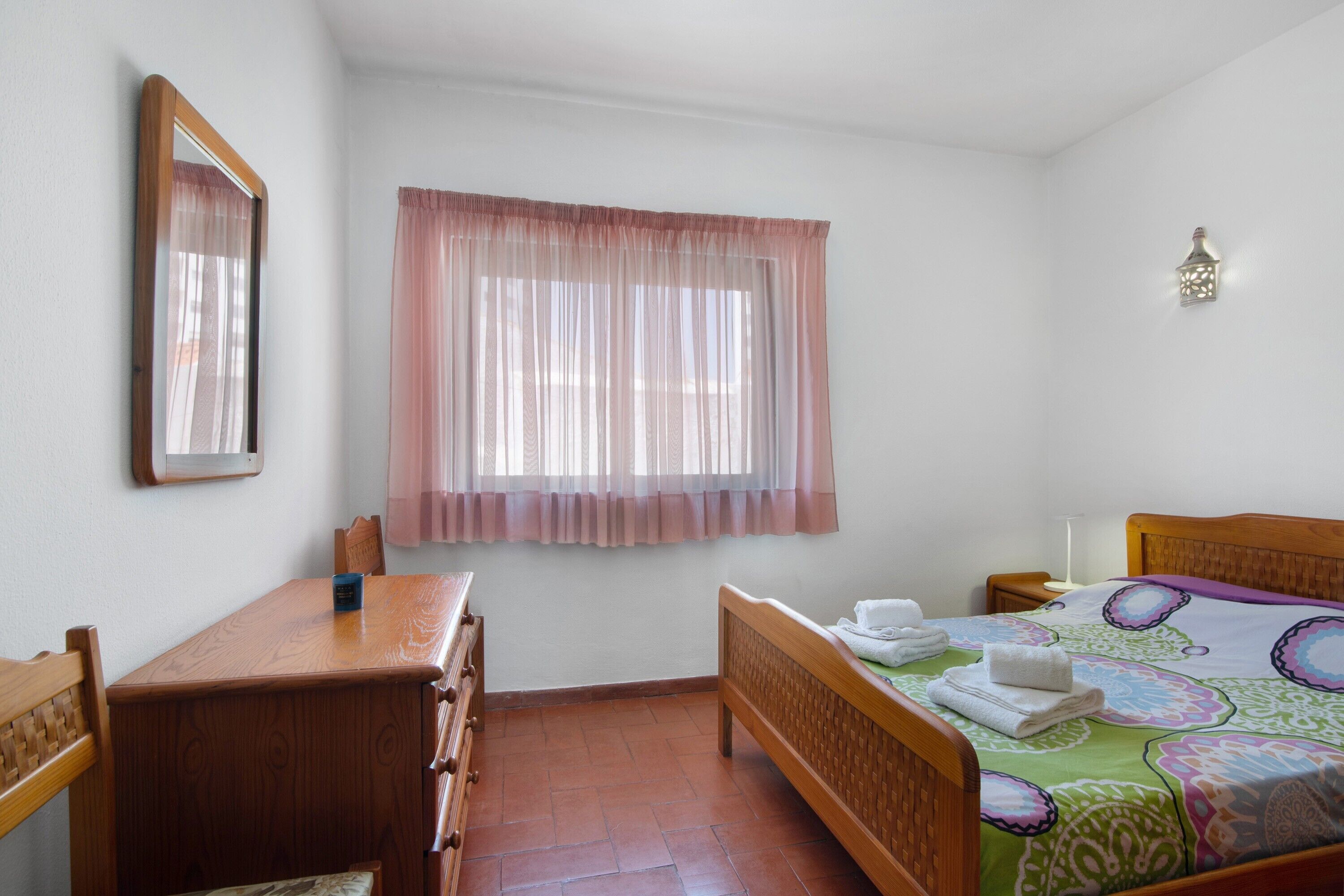 A bedroom with a wooden bed, a bedside table, a chair, a window with sheer curtains, and a wall-mounted lamp.
