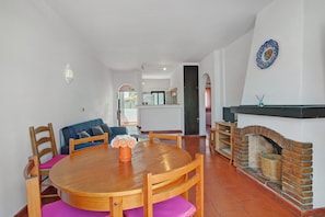 Un coin repas avec une table ronde en bois, des chaises, un vase de fleurs, une cheminée en briques et, au fond, une cuisine.