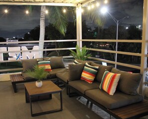 Cómoda zona de estar al aire libre con un sofá y una mesa, rodeada de grandes ventanales con vistas al cielo nocturno y palmeras.