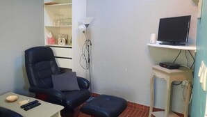 Una habitación pequeña con un sillón de cuero negro, una mesita con un tazón y controles remotos, un televisor en un estante y una lámpara de pared.