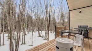 A deck with a grill and chairs, overlooking a snowy forest.