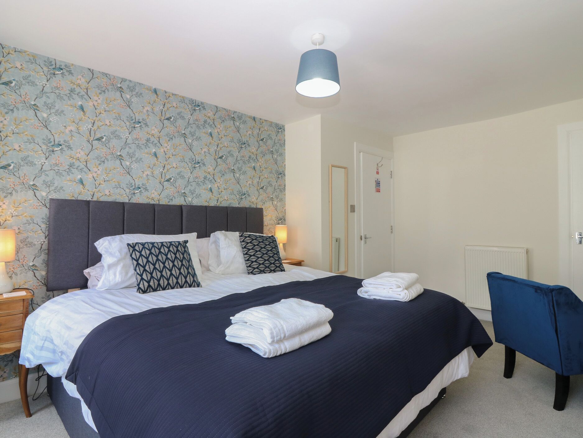 Una cama bien hecha con una colcha azul oscuro y almohadas blancas, un cabecero azul y un estampado floral en la pared.