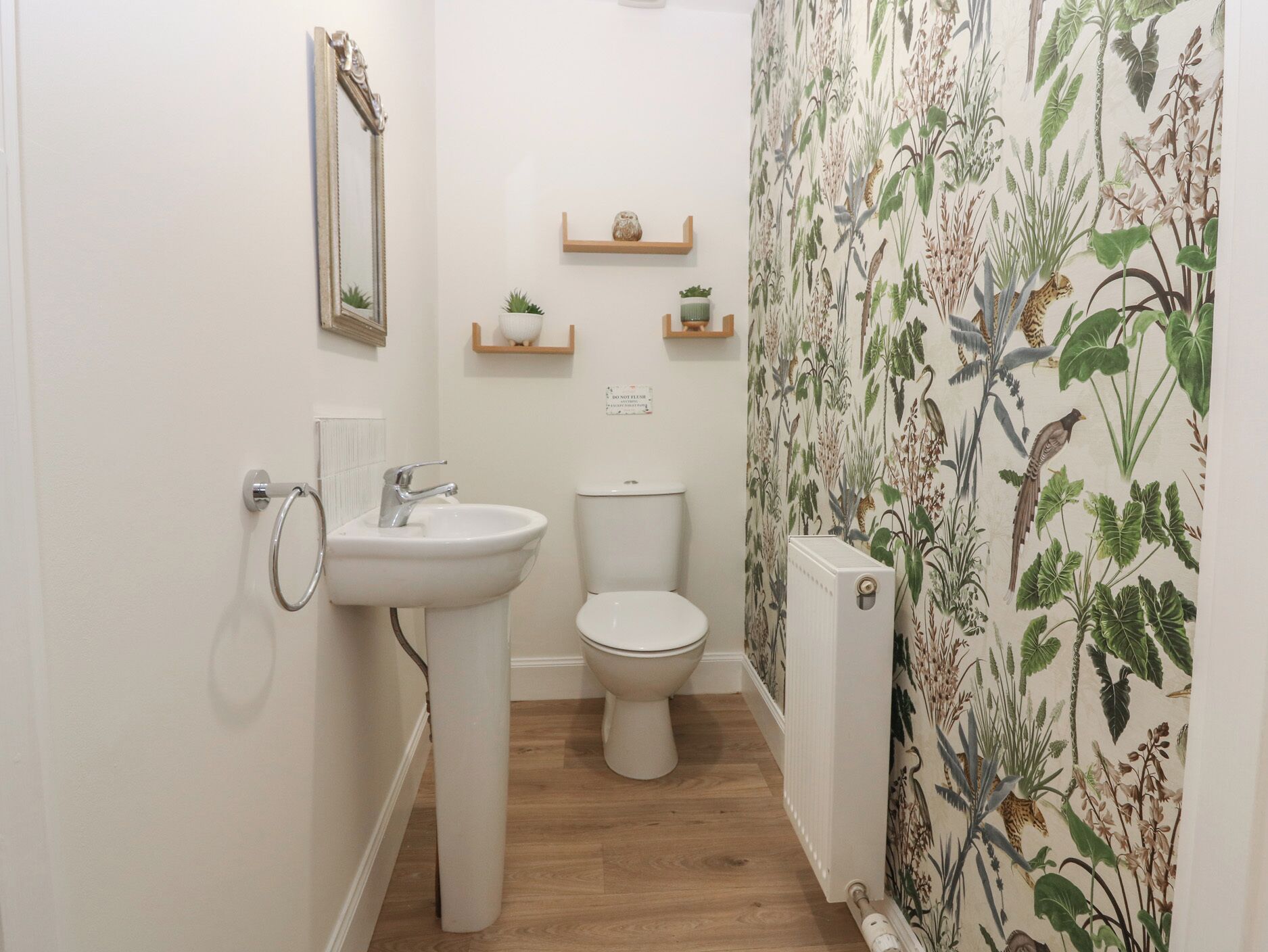 Un baño con lavamanos, inodoro y radiador blancos. Hay dos pequeñas estanterías con objetos decorativos.