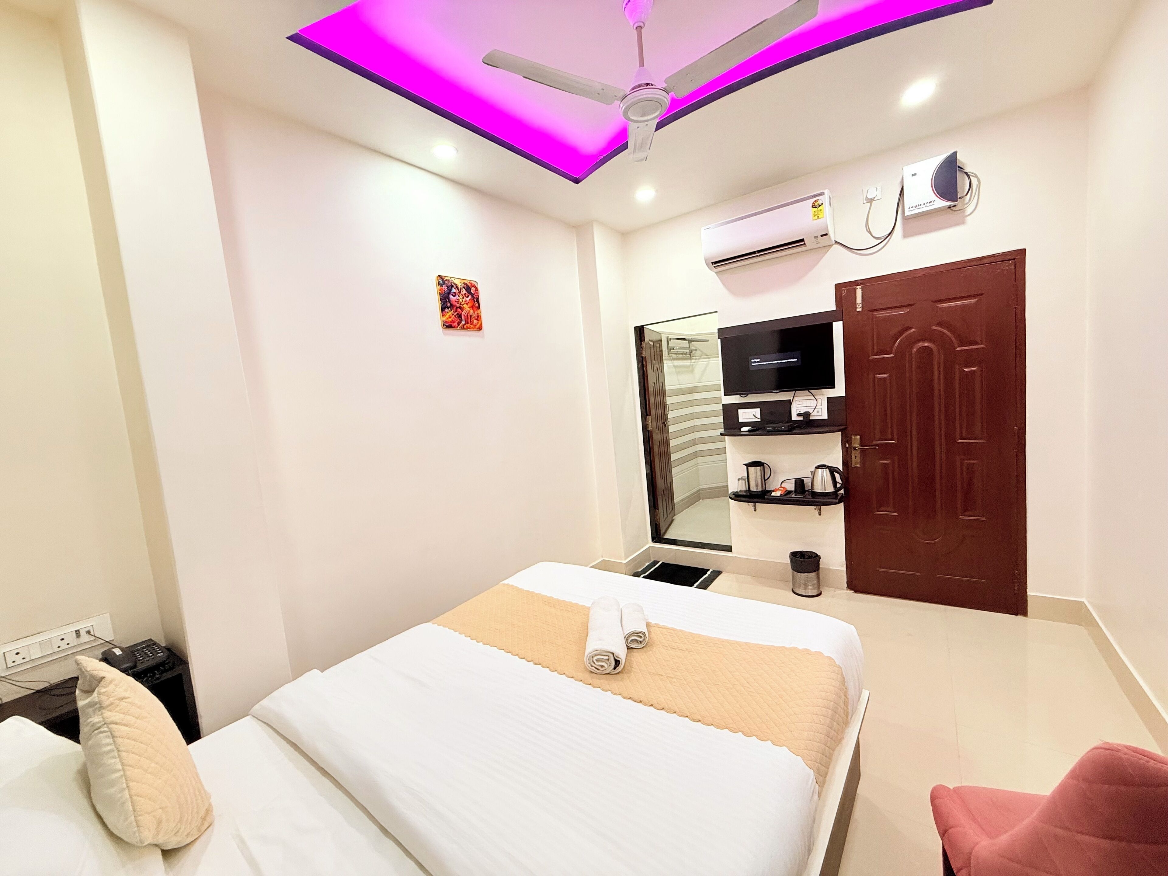 Een hotelkamer met een bed, een televisie, een airconditioningapparaat, een deur en een klein plankje met toiletartikelen.