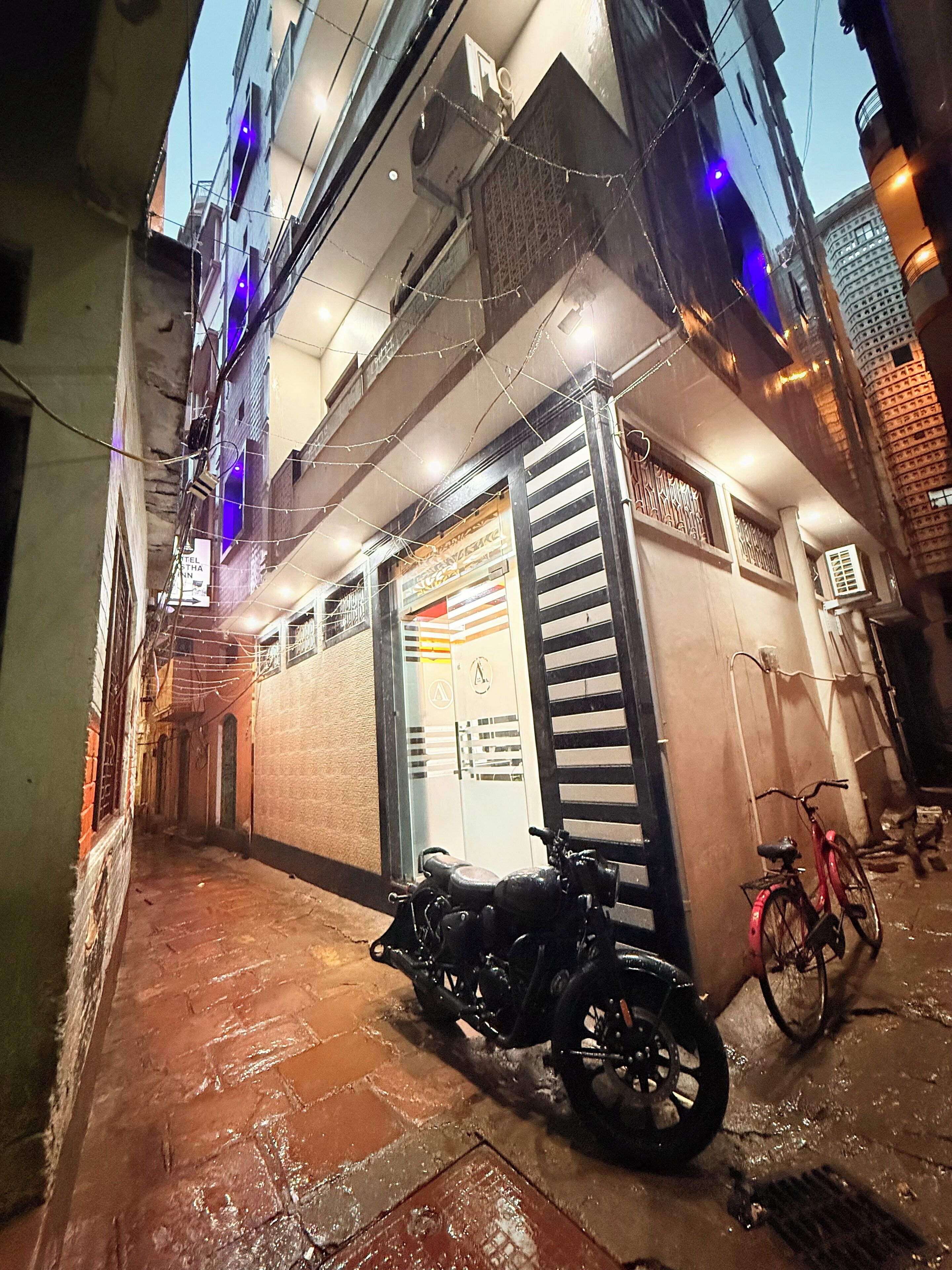 Une ruelle étroite où est garée une moto, devant un immeuble aux fenêtres grillagées et sur lequel on peut voir une enseigne indiquant qu’il s’agit d’un hôtel.
