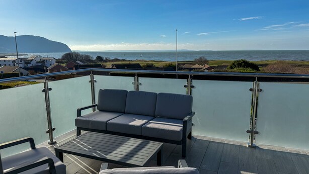 Un balcon doté d’une balustrade en verre, d’un canapé gris, d’une petite table et d’un banc rembourré.