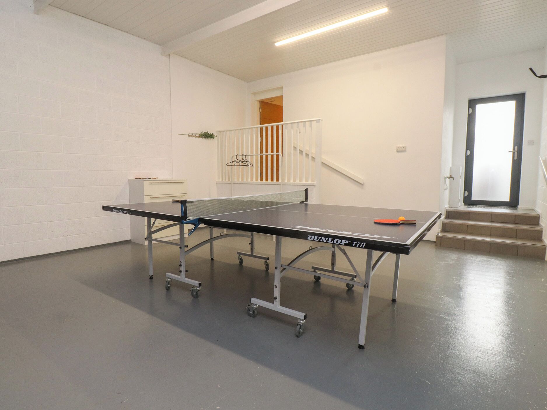 A ping pong table in a room with white walls, a staircase, and a door.