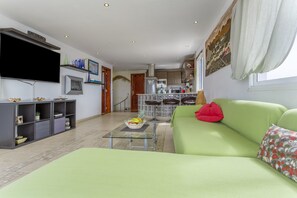 Una sala de estar con un sofá verde, un televisor de pantalla plana, una mesa de centro de vidrio y una cocina al fondo.