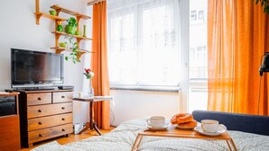 Una habitación acogedora con un aparador de madera, un televisor, una mesita con una bandeja de pasteles y una ventana con cortinas naranjas.
