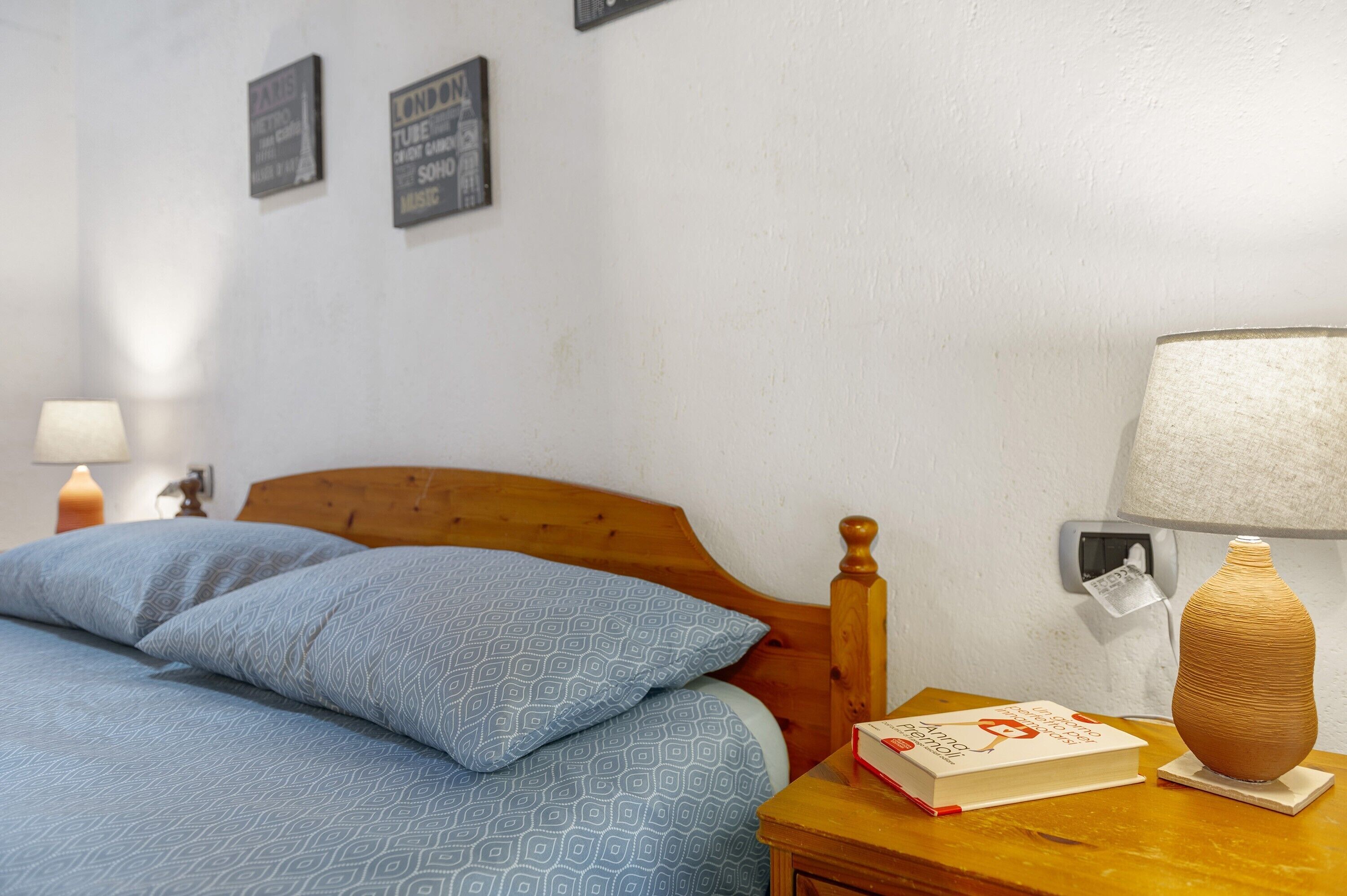 Ein ordentlich bezogenes Bett mit blauem Muster, ein Holz-Kopfbrett, ein Nachttisch mit Buch und Vase sowie ein an der Wand befestigtes Kunstwerk.