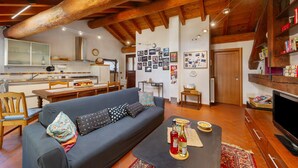 A cozy living room with a sofa, coffee table, and TV, wooden beams on the ceiling, and a kitchen in the background. - 'The Traveler’s Attic' Apartment with Mountain View, Private Terrace and Wi-Fi (Pont Saint Martin)