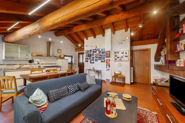 A cozy living room with a sofa, coffee table, and TV, wooden beams on the ceiling, and a kitchen in the background.