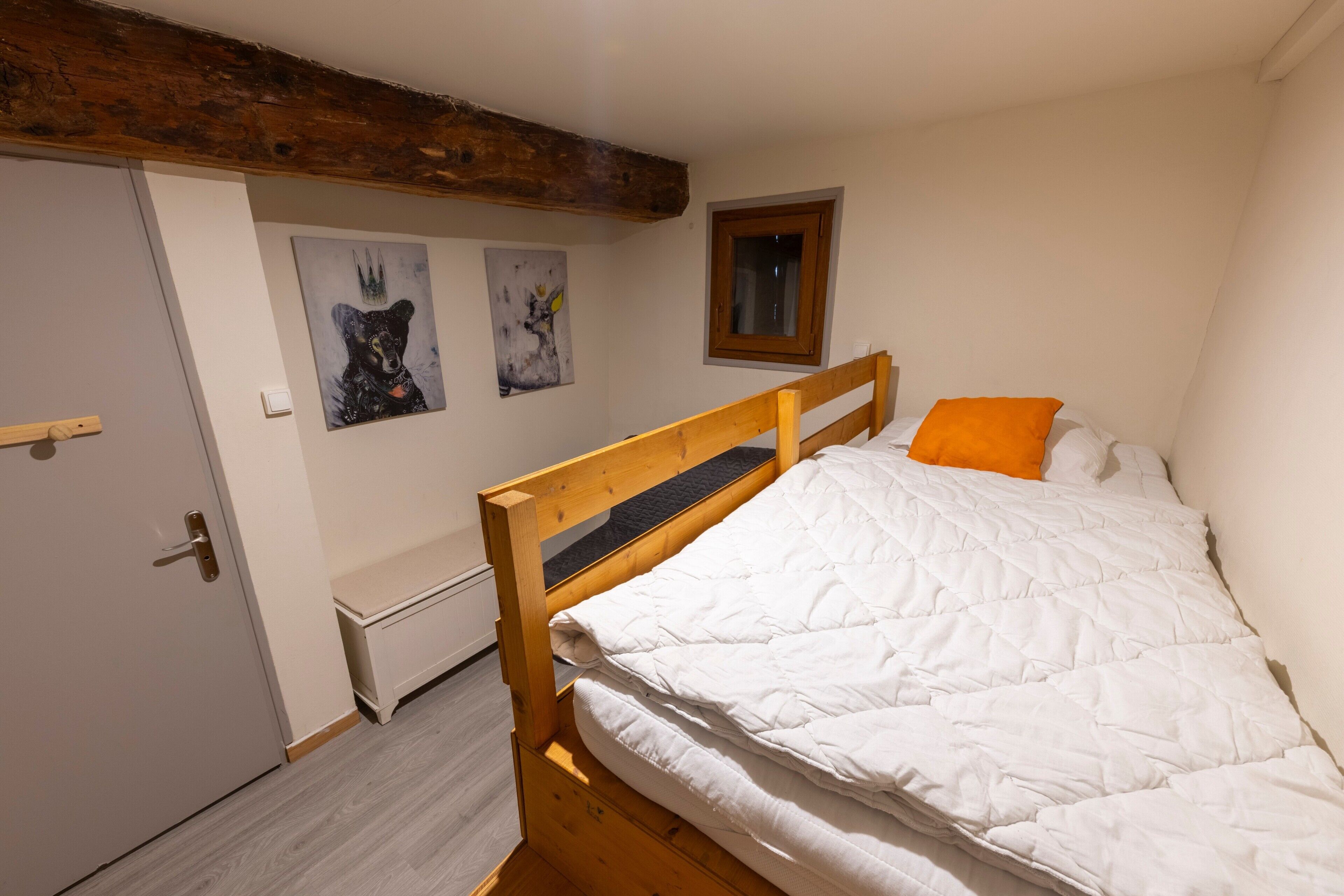 A bedroom with a wooden bed, white bedding, a small window, and artwork on the wall.