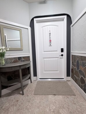 A white door with a 'WELCOME' sign, a mirror, a vase with flowers, and a stone wall. - Resort studio apartment with private hot tub, fire pit, gym, and access to pool. (Ellicott City)