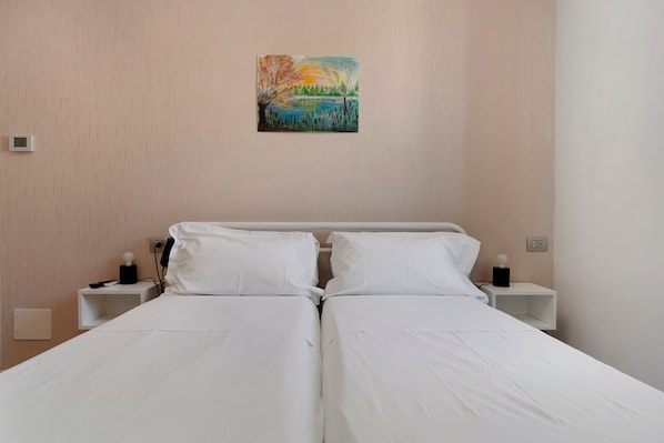 Una cama doble con sábanas blancas, dos mesitas de noche con lámparas, un cuadro en la pared y un interruptor de luz.