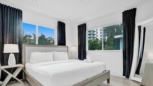A modern hotel room with a large bed, two bedside tables, a nightstand, and a view of the cityscape through large windows.