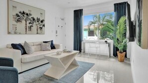A modern living room with a white sofa, a coffee table, and a dining area with a glass table and chairs. There is a large window with curtains, a potted plant, and a painting on the wall.