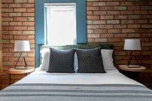A hotel room with a brick wall, a window with blinds, a bed with two pillows, and two bedside tables with lamps.