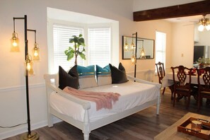 A bedroom with a white bed, a large window with blinds, a potted plant, and a dining area with wooden chairs and a table.