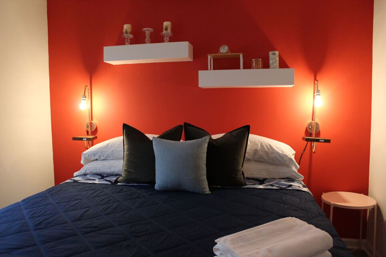 Un lit recouvert d’une couette bleu foncé, avec trois oreillers et une serviette blanche posée sur le côté. Au-dessus du lit, il y a deux étagères fixées au mur, chacune ornée d’une petite horloge et d’objets décoratifs.