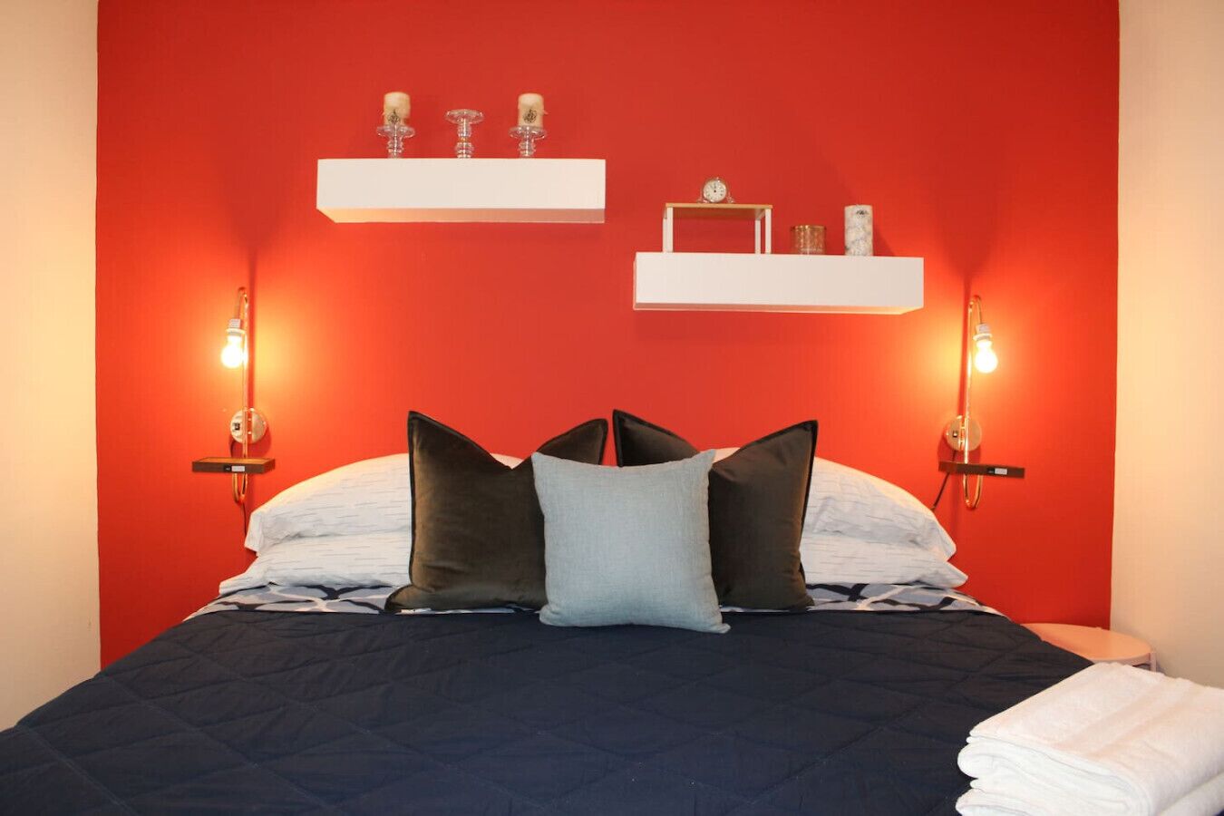Una cama con un edredón oscuro, almohadas blancas y una sola almohada de color claro. Hay dos estantes montados en la pared encima de la cama, uno con tres velas y otro con un reloj y una vela.