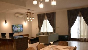 A modern living room with a dining area, featuring a wooden dining table, comfortable seating, a TV, and a unique ceiling light fixture. - Golden Square Resort (Riyadh)