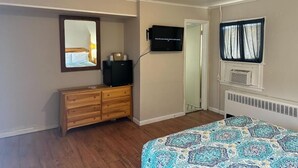 Une chambre avec une commode en bois, un lit avec une literie à motifs, un téléviseur fixé au mur et une fenêtre avec des rideaux.