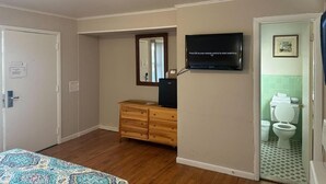 Une chambre d’hôtel avec une commode en bois, un téléviseur fixé au mur, une porte donnant sur une salle de bain équipée de toilettes et d’un lavabo, et une couvre-lit à motifs.