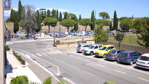 Un parking avec plusieurs voitures, un ciel bleu dégagé et, au fond, un mélange d’arbres et de bâtiments.