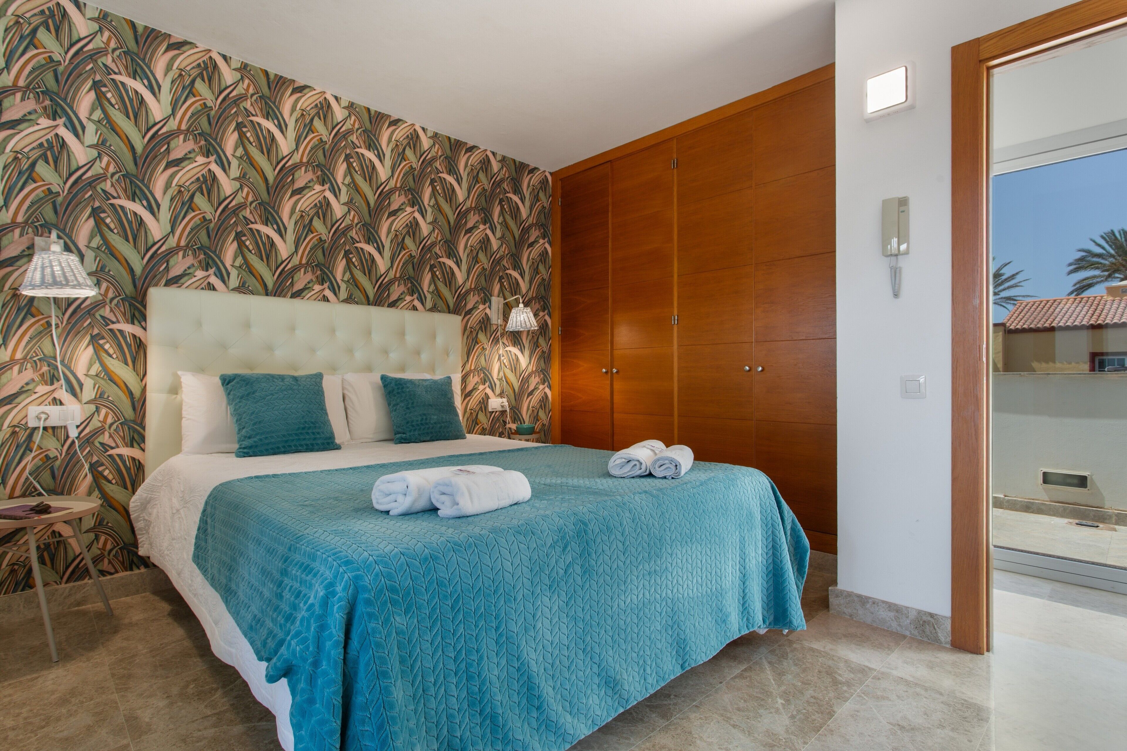 A bedroom with a bed, a wooden wardrobe, a nightstand, and a wall with a leaf pattern.