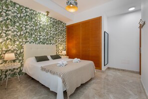 A bedroom with a bed, a wooden wardrobe, a wall with a leafy pattern, and a round table with a lamp.