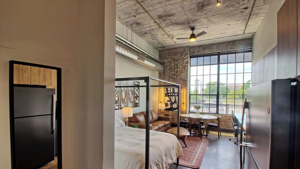 Habitación de hotel compacta con cama, zona de estar con mesa y sillas, refrigerador y un ventanal con vistas al exterior.