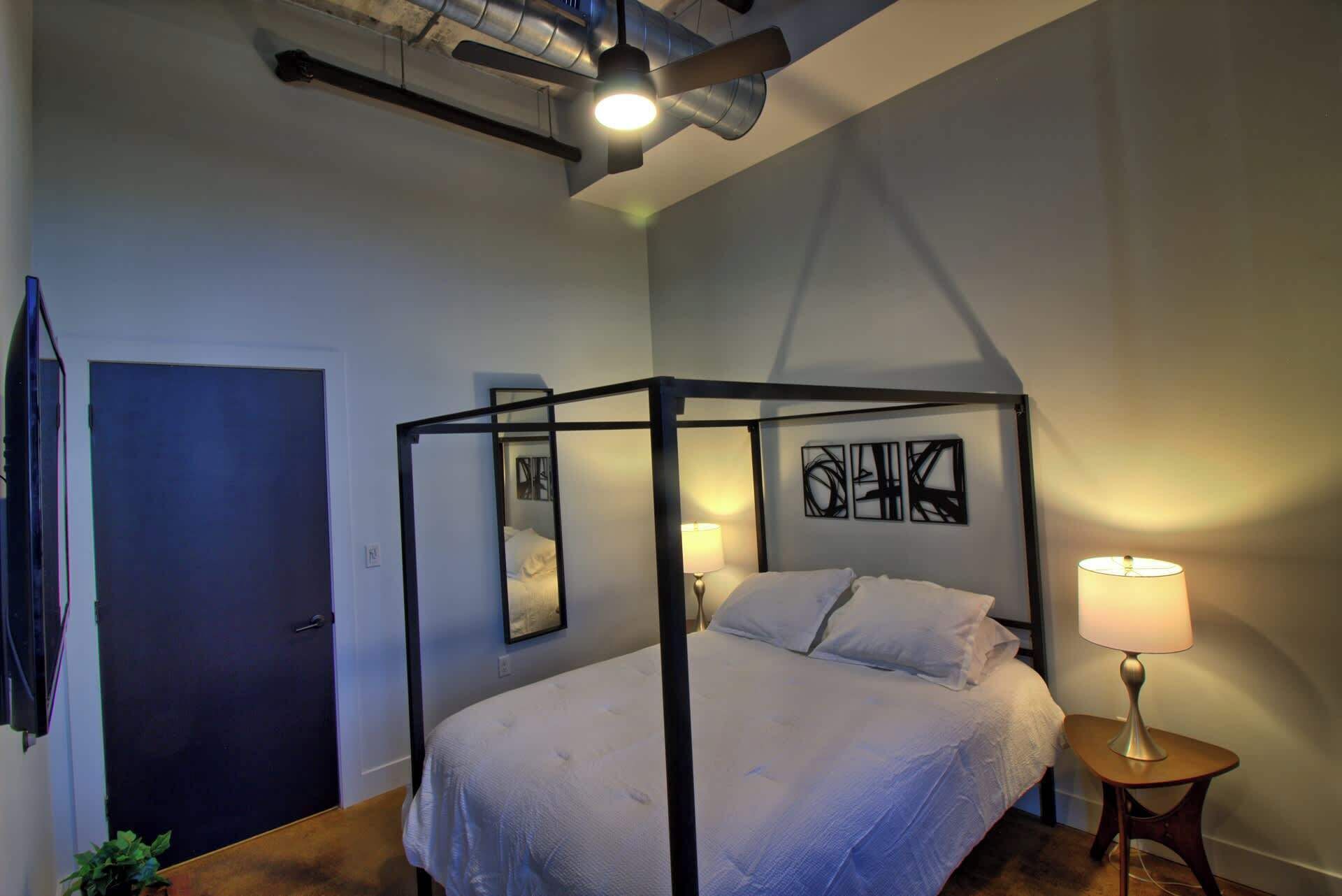 A modern bedroom with a four-poster bed, a bedside table with a lamp, and a ceiling fan.