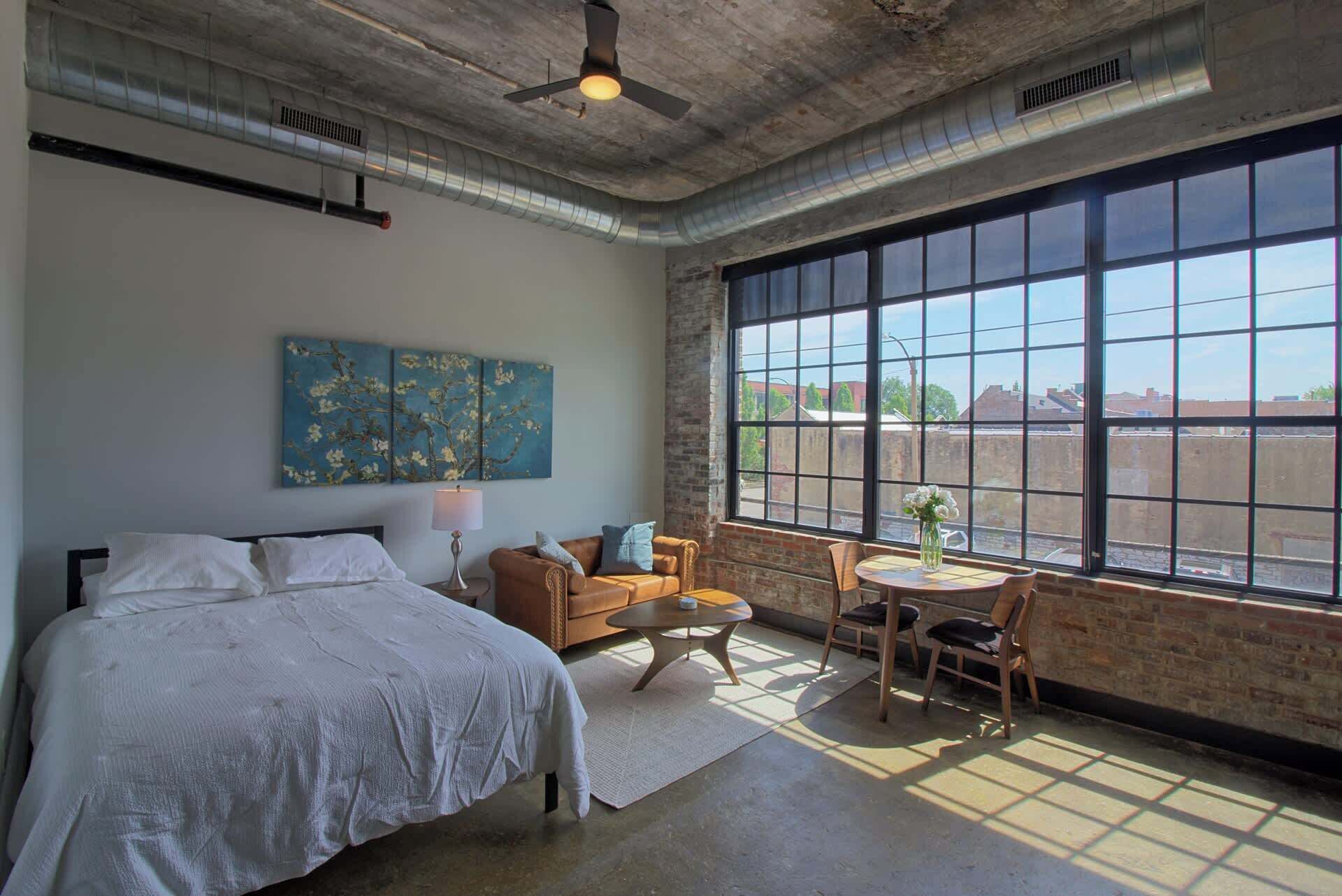 A bedroom with a large window, a bed, a sofa, a small table, and a chair.