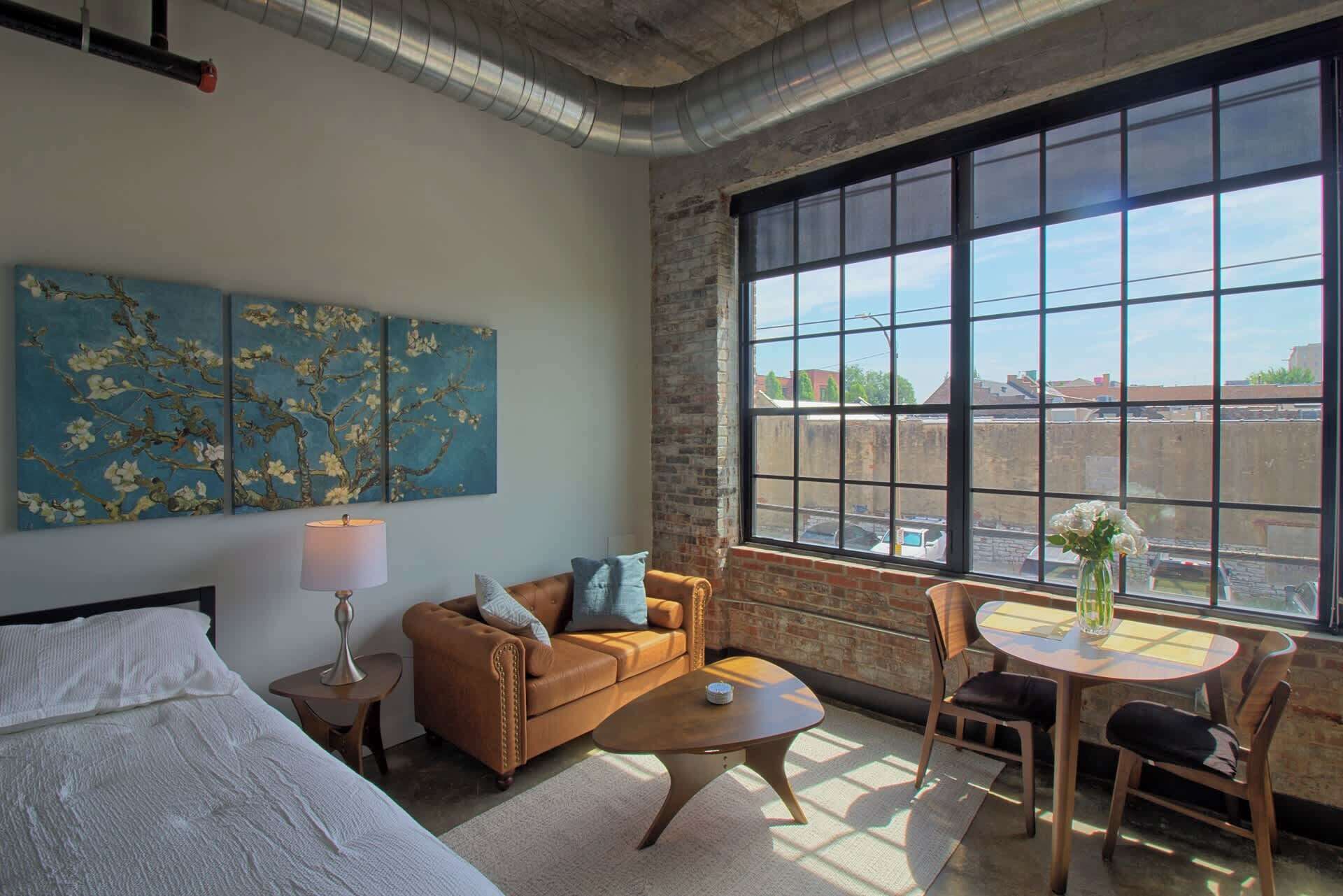 A modern hotel room with a large window, a bed, a sofa, a coffee table, and a small table with a vase.