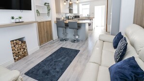 Un salon moderne avec un canapé, une table basse et un téléviseur fixé au mur. On y trouve également un coin cuisine avec une table à manger et des chaises, ainsi qu’un petit îlot avec des tabourets de bar.
