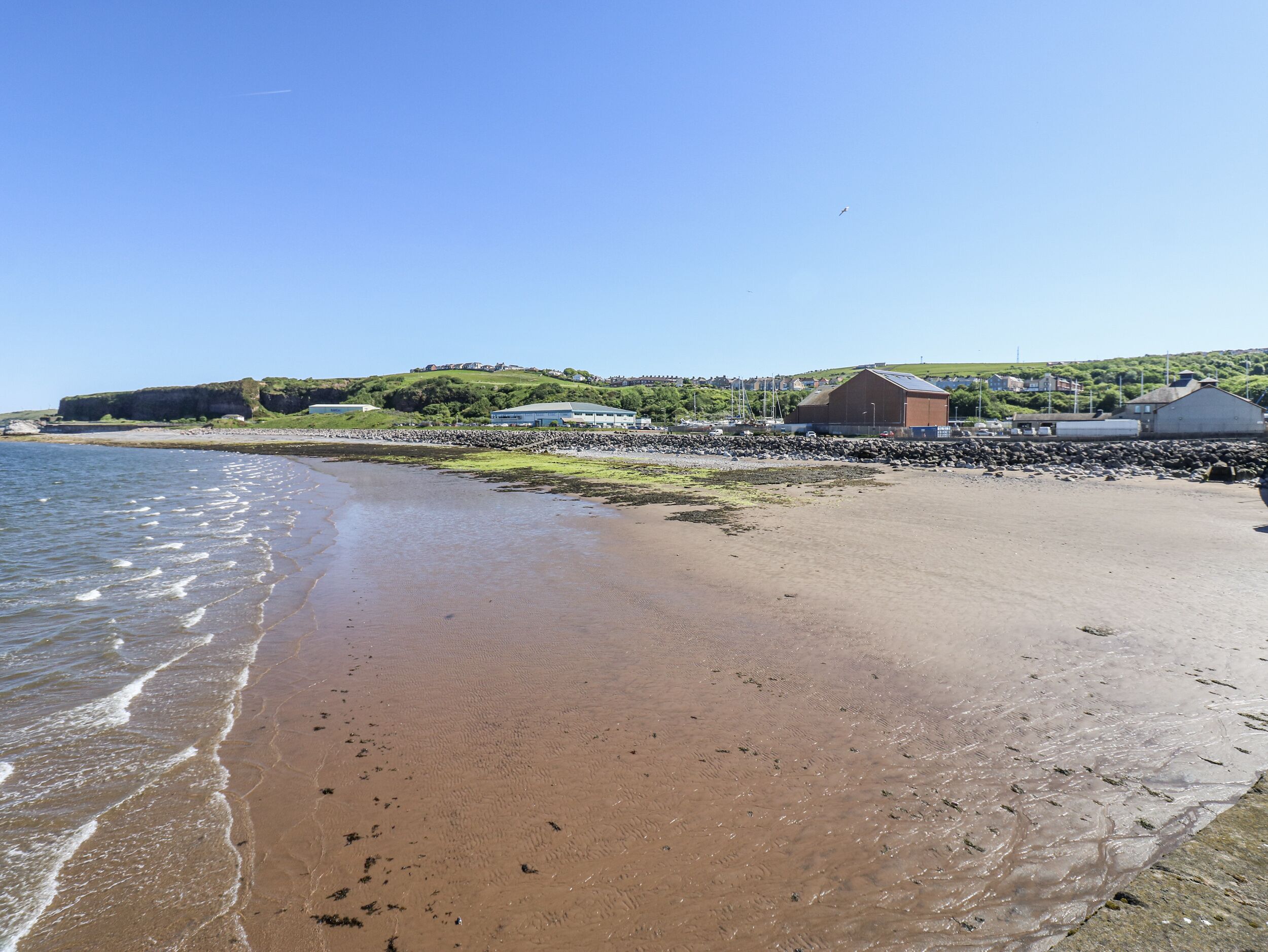 A beach with a rocky shoreline, a building with a flat roof, and a grassy hill in the background.
