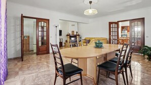 A dining area with a round table, four chairs, a chandelier, and a kitchen in the background.