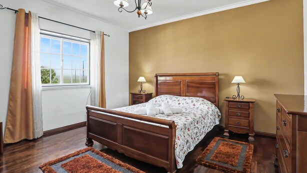 A bedroom with a wooden bed, two bedside tables with lamps, a chandelier, and a view through a window with curtains.