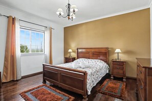 A bedroom with a wooden bed, two bedside tables with lamps, a chandelier, and a view through a window with curtains.