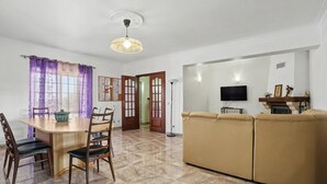 A living room with a beige sofa, a dining table with chairs, a TV mounted on the wall, and a ceiling light fixture.