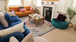 A cozy living room with a sofa, armchair, and a small table with cups and a teapot. There is a fireplace with a mirror above it and a potted plant in the corner. - Hampton House (St Bees)