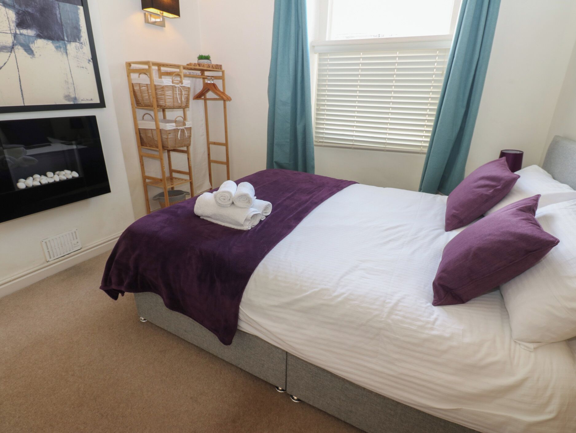 A neatly made bed with a purple blanket and pillows, a wooden side table with baskets, a wall-mounted TV, and a window with blinds.