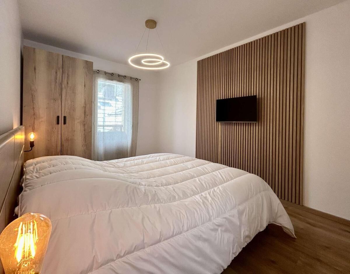 A modern bedroom with a large bed, a flat-screen TV mounted on a wooden panel wall, and a pendant light above the bed.