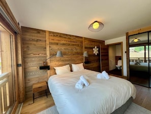 A bedroom with a wooden wall panel, a large bed with white linens, two bedside lamps, and a view through a glass door. - Wifi, 50m from resort center, 2nd floor, view resort, balcony, tv, ski locker, 98m², Les Gets (Les Gets)