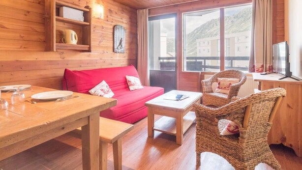 Una stanza accogliente con pareti in legno, un divano rosso, un tavolo da pranzo, sedie in vimini e un'ampia finestra con vista su edifici e colline.