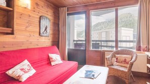 Interno di una baita in legno con un divano rosso, una poltrona in vimini e un'ampia finestra che si affaccia su edifici e colline.