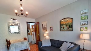 A living room with a dark blue sofa, a small table with a white tablecloth, a chandelier, and framed pictures on the walls. - 100m from ski slopes, 1st floor, view village, balcony, tv, 26m², Le Grand Bornand (Le Grand-Bornand)