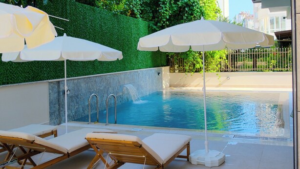 A poolside area with lounge chairs and umbrellas, a green hedge, and a clear pool. - Solmare Suites (Konyaalti)