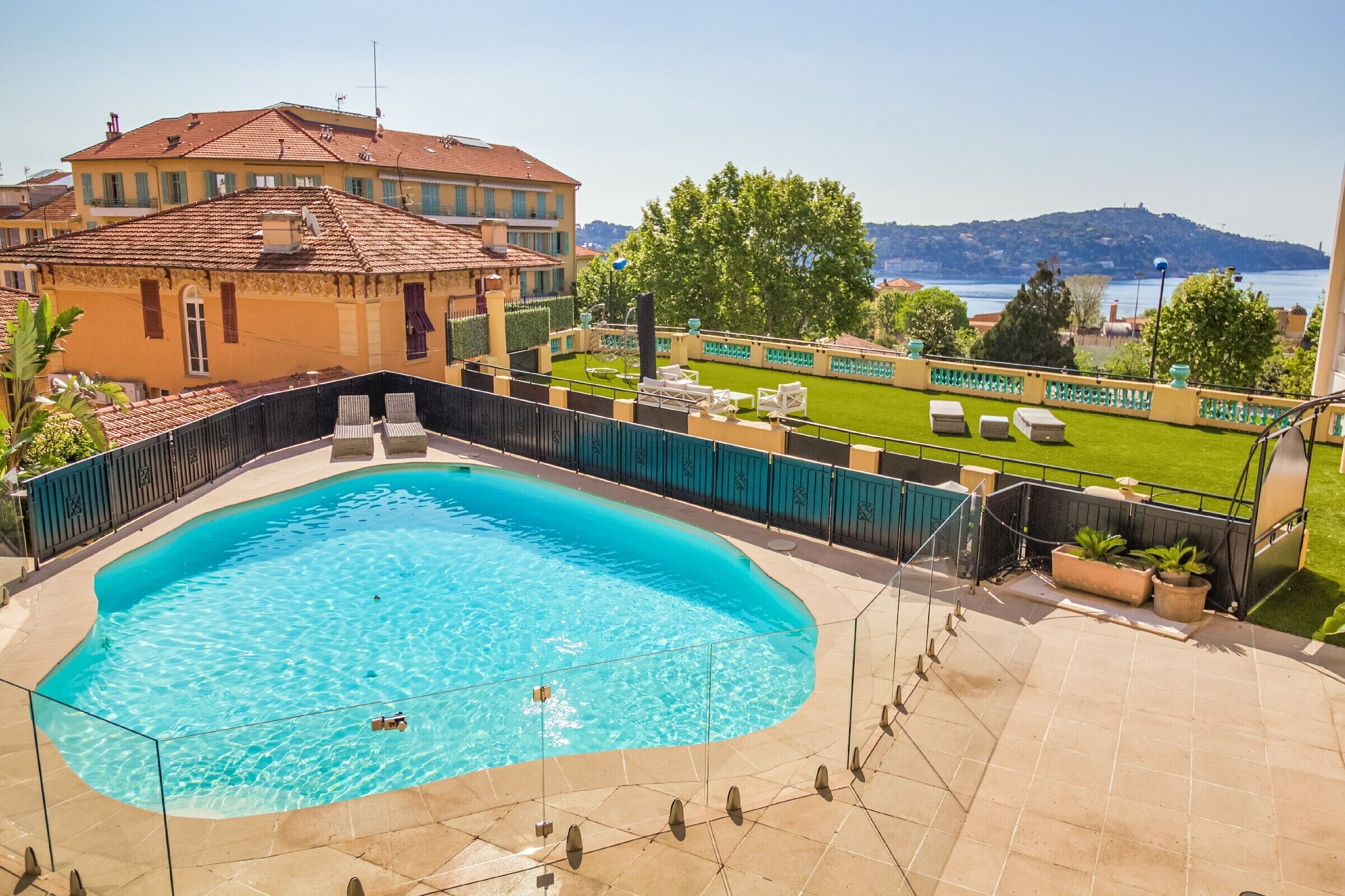 Une piscine à l’eau d’un bleu limpide, entourée d’une terrasse carrelée, avec en arrière-plan une colline et un plan d’eau.